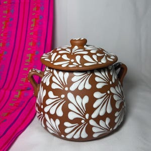 May include: A brown ceramic pot with a lid, adorned with white floral patterns. The pot has two small handles and a matching knob on the lid. The background includes a pink textile with a striped design.