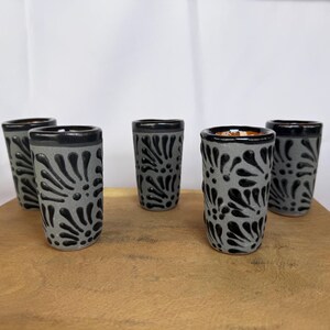 May include: Five gray ceramic shot glasses with black floral designs. The shot glasses have a black rim and are arranged on a wooden surface. The design is a repeating pattern of stylized leaves.