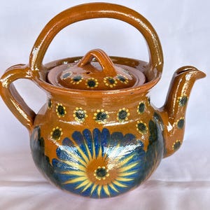 May include: A brown glazed ceramic teapot featuring a large blue and yellow floral design on the front. The teapot has a rounded body, a curved handle, a matching lid with a small handle, and a spout decorated with small yellow flower accents.