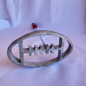 May include: A cast iron branding iron in the shape of a football. The iron has a handle with a red grip. The branding iron is silver and has a detailed design. The background is a white sheet.