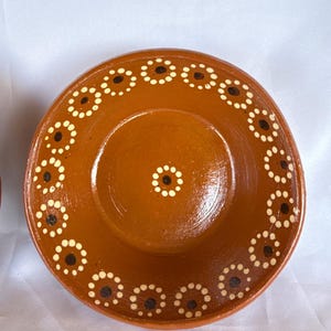 May include: A round, terracotta-colored ceramic bowl with a glossy finish. The interior features a hand-painted pattern of small, white-dotted circles surrounding a dark brown center, arranged in a ring along the rim and a single dot in the middle.