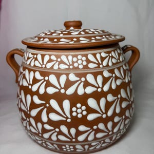 May include: A brown ceramic pot with a lid, adorned with white floral patterns. The pot has two handles and a knob on the lid. The design is repeated around the pot's body and on the lid, creating a cohesive look.