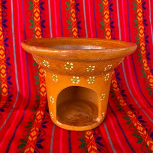 May include: A handcrafted, terracotta-colored pottery incense burner with a wide, flared rim and a cylindrical base. The base features a large opening and is decorated with white and green floral accents. The burner sits on a vibrant red textile background.
