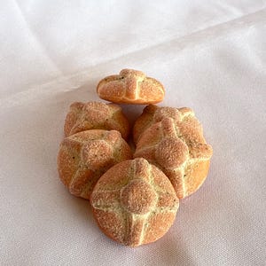 May include: A collection of five small, round, golden-brown baked goods, each featuring a central circular piece and four radiating strips of dough on top, creating a cross-like pattern with a textured, granular surface finish.