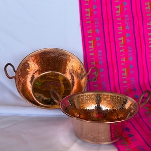 May include: Two round, hammered copper bowls with small, curved side handles. The bowls feature a polished, reflective interior surface and a textured exterior finish, showcasing the metallic orange-brown color of the copper material.