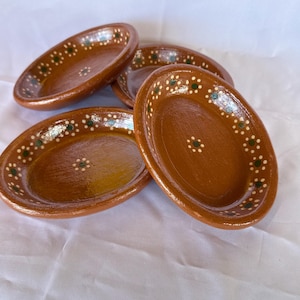 May include: A set of four oval-shaped brown clay bowls with a glossy finish. Each bowl features a decorative rim and center pattern consisting of small white dots surrounding a single green dot, creating a repeating floral-like motif around the edge.