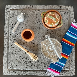 May include: A gray stone slab with a variety of kitchen tools, including a metal tortilla press, a small clay pot, a metal strainer, a wooden stirring utensil, and a small clay pot with a lid. A colorful Mexican blanket is also on the slab.