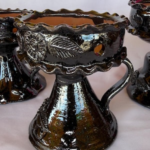 May include: A dark brown, glossy ceramic incense burner with a scalloped rim and a flared base. The exterior features embossed floral and leaf motifs, a single handle on the side, and small circular cutouts around the upper bowl section.