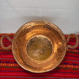 May include: A hammered copper bowl with two wooden handles. The bowl has a textured surface and a golden hue. The bowl is sitting on a red and multi-colored striped textile.