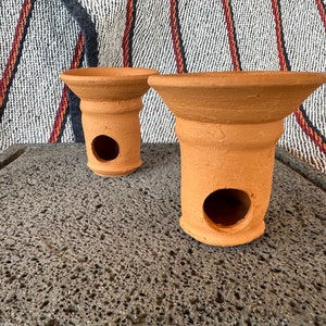May include: Two terracotta incense burners with a hole in the bottom. The burners are on a dark gray textured surface.