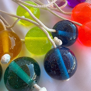 May include: A collection of colorful glass balls with glitter and a white string attached. The balls are in various colors, including yellow, green, blue, orange, and purple. The balls have a clear, shiny appearance.