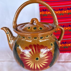 May include: A brown glazed clay teapot featuring a large hand-painted red and yellow flower on the front, dark green leaf accents, and a matching lid with a small handle. The teapot has a curved spout and a high arched handle over the top.