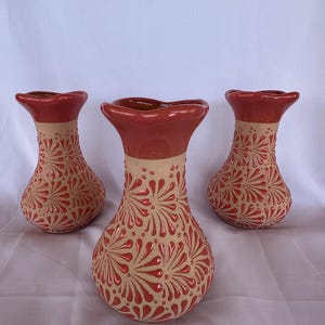 May include: Three ceramic vases with a red and tan color scheme. The vases have a bulbous shape with a flared rim. The tan-colored bodies are decorated with a repeating red floral pattern. The rims are a solid red color.