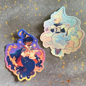 May include: Two die-cut stickers featuring illustrated characters. One sticker shows a dark-haired figure in a blue robe with orange flames and a scroll. The other sticker depicts a light-haired figure in a pale robe surrounded by swirling blue water.
