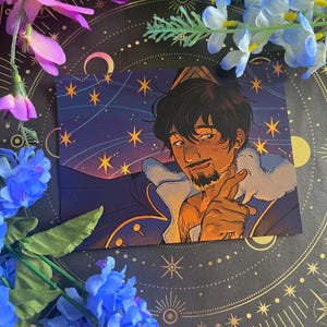 Puede incluir: Una impresión artística que presenta una ilustración estilizada de un hombre con cabello oscuro y barba, señalando. La obra de arte tiene un tema celestial con estrellas y una paleta de colores azul y dorado. El hombre lleva un abrigo azul con cuello de piel blanco.