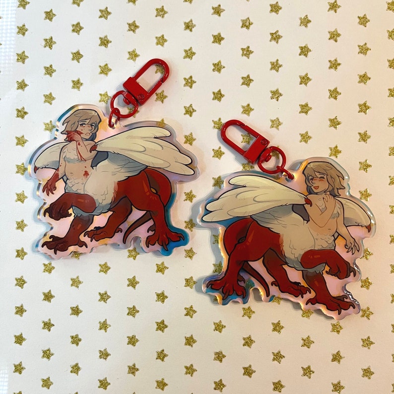 May include: Two red and white keychain charms featuring a winged creature with a human head. The creature is red and white with a white wingspan. The charms are attached to red metal key rings.