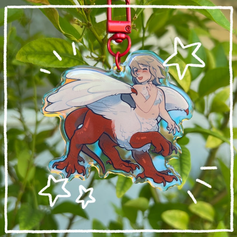 May include: A clear acrylic keychain featuring a cartoon illustration of a red and white griffin with wings. The griffin is smiling and has a human-like face. The keychain is hanging from a red metal chain.