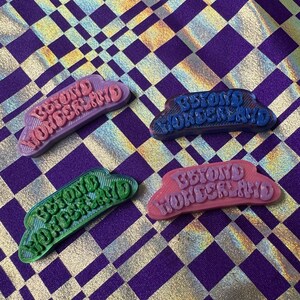 May include: Four colourful 3D printed plastic badges with the text "Beyond Wonderland" in a wavy font. The badges are pink, blue, green, and purple.