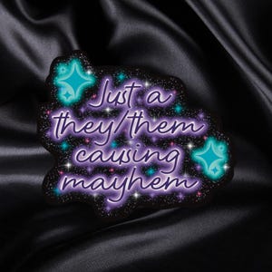 May include: A sticker with a black background and a purple, starry design. The text "Just a they/them causing mayhem" is written in a whimsical font. Teal starbursts accent the design.