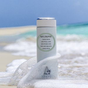 May include: White water bottle with a black circle containing the text "MURPH" and the workout routine: "1 MILE RUN, 100 PULL-UPS, 200 PUSH-UPS, 300 AIR SQUATS, 1 MILE RUN". The bottle is partially submerged in white foam on a beach.