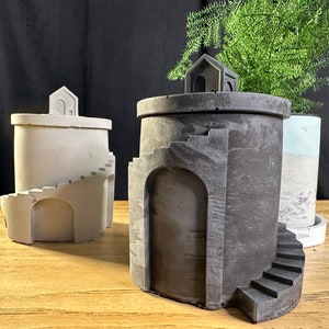 May include: Three concrete planters with a unique design featuring a small house on top and a spiral staircase on the side. The planters are in different shades of gray and white.