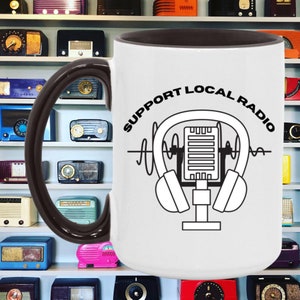 May include: White ceramic mug with a brown handle and interior. The mug features a black and white graphic of a microphone and headphones with the text "SUPPORT LOCAL RADIO" in a circular design. The background shows a collection of vintage radios.