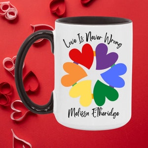 May include: White ceramic coffee mug with a black handle and rim. The mug has a rainbow heart design with the text "Love Is Never Wrong" and "Melissa Etheridge" printed on it.