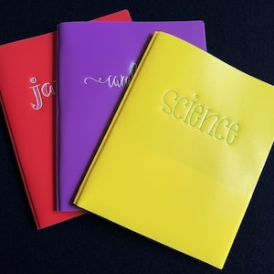 Puede incluir: Tres carpetas rectangulares en rojo, morado y amarillo. La carpeta roja tiene el texto "ja" bordado en blanco. La carpeta morada presenta el texto "caro" en blanco. La carpeta amarilla muestra la palabra "science" cosida en verde.
