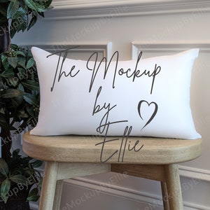 以下が含まれることがあります： 「The Mockup by Ellie」という文字と小さなハートマークが書かれた白い枕。枕は木の踏み台の上に置かれています。