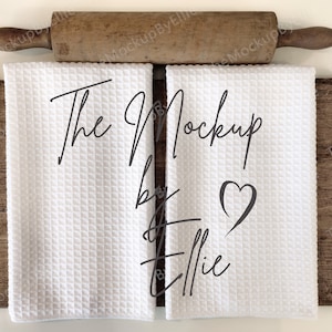 May include: Two white kitchen towels with the text "The Mockup by Ellie" and a heart symbol on a wooden surface with a rolling pin on top.