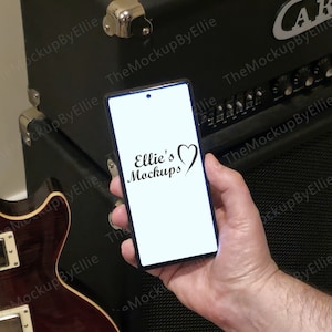 May include: A black smartphone with a white screen displaying the text "Ellie's Mockups" with a heart icon. The phone is being held in a person's hand in front of a black guitar amplifier.