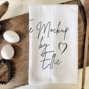 May include: White kitchen towel with the text "The Mockup by Ellie" and a small heart design. The towel is on a wooden cutting board with a rolling pin, two eggs, and a wooden spoon.