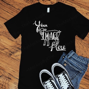 May include: Black t-shirt with a white goat graphic and the text "Your Image Here". The shirt is laying on a wooden surface with a pair of black sneakers and blue jeans.