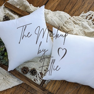 Könnte beinhalten: Zwei weiße Kissen mit dem Text "The Mockup by Allie" und einem Herzsymbol auf einem Kissen. Die Kissen liegen auf einer Holzoberfläche mit einer braunen Decke.
