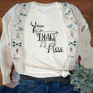 May include: Cream-colored t-shirt with the text "Your Image Here" and a graphic of a lamb. A beige cardigan with floral embroidery on the sleeves is draped over the shirt. The shirt is paired with blue jeans and a brown belt.
