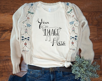 May include: Cream-colored t-shirt with the text "Your Image Here" and a graphic of a lamb. A beige cardigan with floral embroidery on the sleeves is draped over the shirt. The shirt is paired with blue jeans and a brown belt.