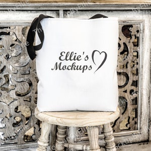 以下が含まれることがあります： 黒い持ち手付きの白いキャンバス地のトートバッグ。バッグには「Ellie's Mockups」という文字とハートのデザインがプリントされています。バッグは木製の踏み台の上に置かれています。