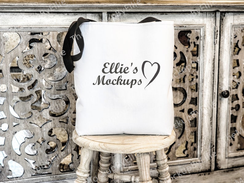以下が含まれることがあります： 黒い持ち手付きの白いキャンバス地のトートバッグ。バッグには「Ellie's Mockups」という文字とハートのデザインがプリントされています。バッグは木製の踏み台の上に置かれています。