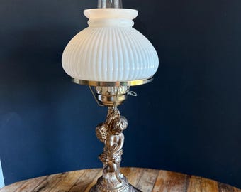 Könnte beinhalten: Ein weißer Glasschirm mit einer goldenen Basis, die eine Cherubinskulptur zeigt. Der Lampenschirm hat vertikale Rippen und einen goldenen Rand.