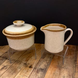 Könnte beinhalten: Ein Vintage-Zuckertopf mit Deckel und ein Sahnekännchen. Beide Teile sind beige gesprenkelt mit braunem Rand. Der Zuckertopf ist rund mit Deckel und kleinem Knauf. Die Sahnekanne hat einen Henkel und einen Ausguss.