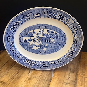 May include: An oval blue and white porcelain serving platter with a traditional willow pattern design. The platter features a blue and white landscape scene with a bridge, trees, and a pagoda.