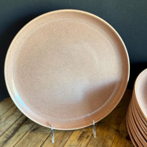 Könnte beinhalten: Ein Set rosa gesprenkelter Keramik-Dinnerteller. Die Teller haben einen leicht erhöhten Rand und eine glatte, matte Oberfläche.