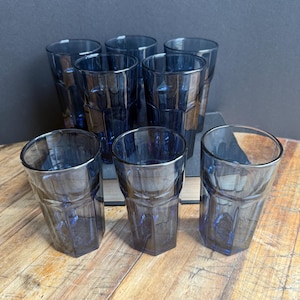 Op de afbeelding: Set van zes blauw getinte drinkglazen. Elk glas heeft een facetontwerp en een cilindrische vorm. De glazen zijn gerangschikt op een houten oppervlak, met enkele glazen op de achtergrond en andere op de voorgrond.