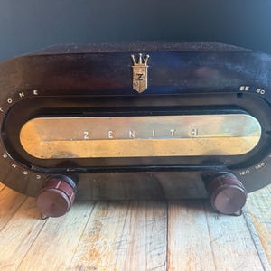 May include: A vintage Zenith radio with a dark brown, oval-shaped plastic casing. The front features a horizontal gold-toned metal panel with the word ZENITH printed in the center. A gold crest logo sits above the dial, which displays numbers 55 to 160.