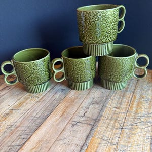 Può includere: Quattro tazze in ceramica verde oliva con un motivo floreale in rilievo sulla parte superiore e scanalature verticali sulla base. Ogni tazza presenta un manico distintivo con due anelli circolari sovrapposti. Le tazze sono disposte in gruppo.