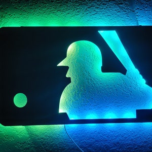 May include: A black metal sign with a green and blue LED light design. The sign features a silhouette of a baseball player with a bat and a baseball. The sign is lit up with green and blue lights.