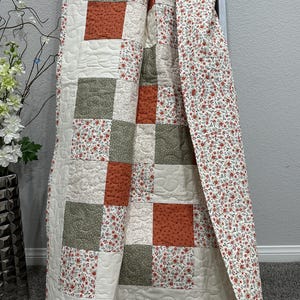 May include: A patchwork quilt featuring a repeating pattern of square blocks in solid rust orange, sage green with white dots, and a cream fabric with a small red and green floral print. The quilt has visible machine quilting in a swirling pattern.
