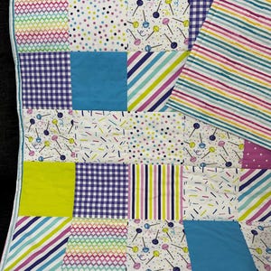 May include: A colorful patchwork quilt with various patterns. The quilt features squares of blue, green, purple, and white fabric with stripes, polka dots, and lollipop designs. The edges are trimmed with a striped border.