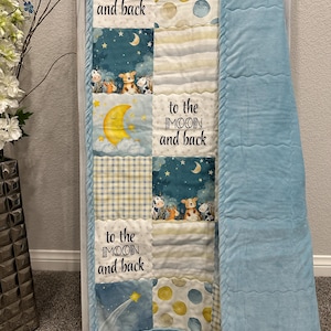 May include: A patchwork baby quilt with the text "to the MOON and back". The quilt features a blue, yellow, and white colour scheme with illustrations of animals, moons, and stars. A solid light blue blanket is attached to the quilt.