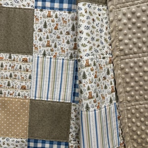May include: A patchwork quilt featuring various fabric patterns. The quilt includes squares of blue gingham, forest animal prints with deer and bears, floral patterns, and solid brown squares. A soft, textured border is visible.